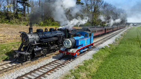 Ronks, Pennsylvania 17 Nisan 2025 - İki klasik buhar lokomotifi paralel raylarda hareket halinde, yeşil zemine doğru dalgalanan beyaz buhar. Bağbozumu demiryolunun cazibesini yakalıyor.