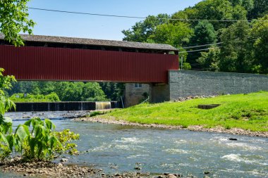 Güneşli bir günde kırmızı kaplı bir köprü, canlı yeşilliklerle çevrili akan bir nehrin üzerine uzanır. Resimli dekor doğa ve tarihi mimariyi birlikte sergiliyor..