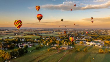 Parlak renkli sıcak hava balonları günbatımında gökyüzünü dolduruyor, kırsal bir bölgede yüzüyorlar. Güneş batarken aşağıda tarlalar ve tarlalar görülebilir, sakin bir atmosfer yaratır..