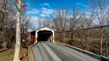 Çıplak ağaçlar ve açık mavi gökyüzüyle çevrili tarihi bir köprü bir yolun kenarında duruyor. Sahne, kırsal kesimde bir kış gününün sükunetini yakalıyor..