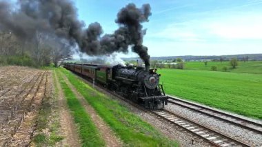 Ronks, Pensilvanya, 18 Nisan 2025 - Klasik bir buhar treni, canlı yeşil alanlar arasında yol boyunca siyah dumanı dalgalandırır. Sahne, klasik ulaşım ve manzaranın çekiciliğini yakalıyor.