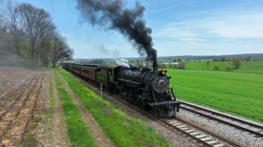 Ronks, Pennsylvania, 18 Nisan 2025 - Büyük bir buharlı lokomotif, açık mavi gökyüzüne kara dumanlar salıyor. Yeşil tarlalar ve ağaçlar buna resim gibi bir zemin hazırlıyor.
