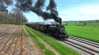 Ronks, Pennsylvania, 18 Nisan 2025 - Pist boyunca uzanan eski bir buhar lokomotifi, açık gökyüzüne siyah duman üflüyor, canlı yeşil alanlar ve tomurcuklanan ağaçlarla çevrili. Sahne