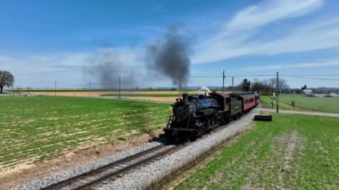 Ronks, Pennsylvania, 18 Nisan 2025 - Pahalı raylar boyunca uzanan tarihi bir buhar treni, canlı yeşil alanlar ve açık mavi bir gökyüzü ile çevrilidir. Motor hareket ederken duman dalgalanır.