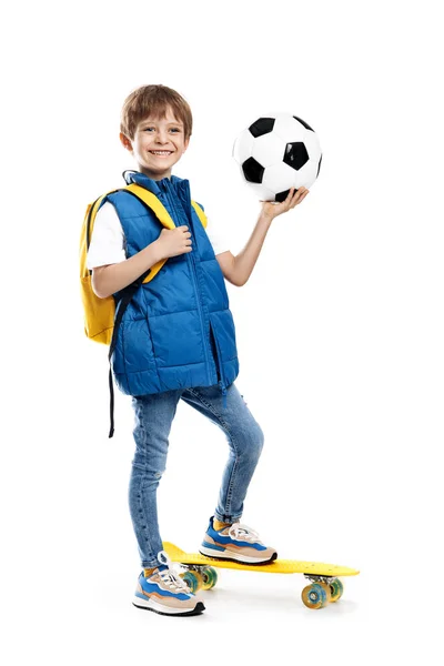 穿着滑板和背包的时髦小男孩拿着白色背景的足球 回学校去 图库图片