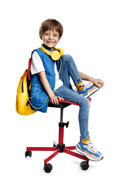 幸せな男の子は白い背景に赤い椅子に座ってヘッドフォンと黄色のバックパックを着て 学校に戻る ストック画像