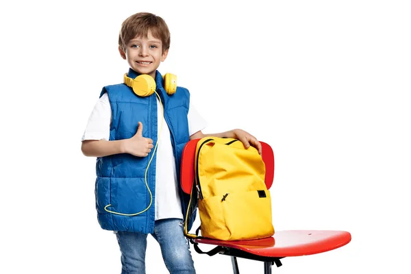 stock image Gleeful little boy with headphone and backpack stay near red chair on white background. Back to School. Show thumb up
