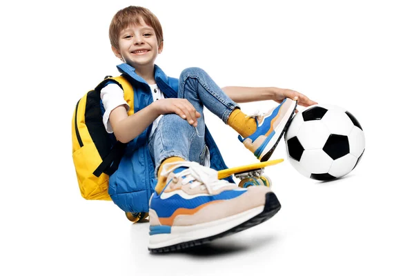 英俊的男孩坐在黄色的滑板上 在白色的背景上抱着腿踢足球 回学校去 图库图片