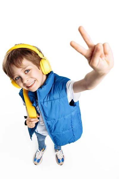笑着的小男孩戴着耳机和黄色的背包 在白色的背景上显示出和平的姿态 回学校去 图库图片