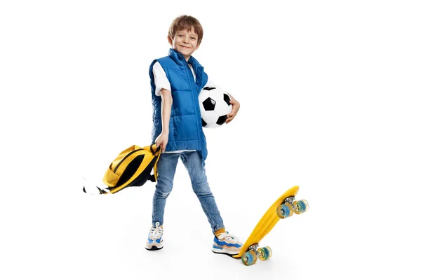 スケートボードとバックパックを白い背景にサッカーボールを保持している小さな男の子のダイナミックなイメージ 学校に戻る ストックフォト