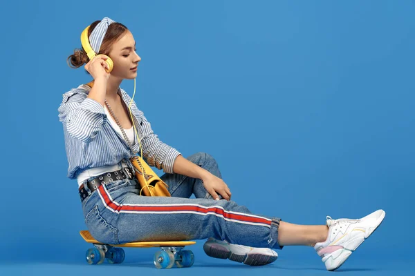 青の背景の上にスケートボードに座っている黄色のヘッドフォンを持つ屈託のない若い女性の完全な長さの写真 ロイヤリティフリーのストック写真