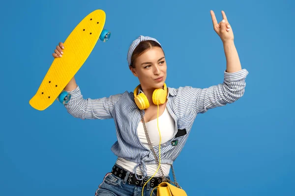 自信を持って若いです女性とともに黄色のヘッドフォンポージングとともにスケートボードショー岩ジェスチャー上の青の背景 ロイヤリティフリーのストック写真