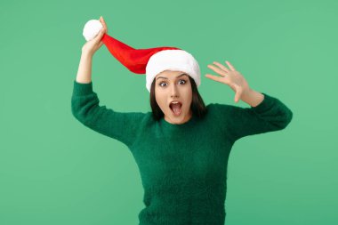 Heyecanlı genç esmer kadın Noel Baba şapkası ve yeşil arka planda yeşil kazak giyiyor.