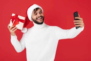 Hippi adam Noel Baba şapkası takıyor ve kırmızı arka planda hediye kutusu tutuyor. Noel ve Yeni Yıl konsepti. Selfie çekin.