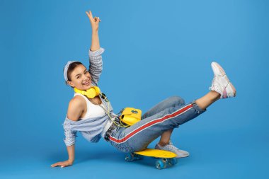 Mavi arka planda kaykayın üzerinde oturan sarı kulaklıklı mutlu, kaygısız genç kadının tam boy fotoğrafı.