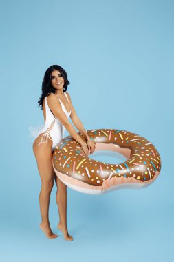 Mayo giymiş çekici genç bir kadın mavi arka planda donut şişirilebilir halkayla mutlu poz veriyor.
