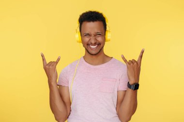 Sarı arka planda rock hareketi gösteren kulaklık takan mutlu Afrikalı Amerikalı genç adam.