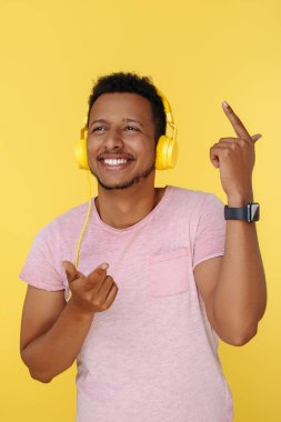 Gülümseyen Afrikalı Amerikalı genç adam kulaklıkla müzik dinliyor ve sarı arka planda dans ediyor. Uzaklara bakıyorum.