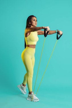 Mavi arka planda geniş bantlı omuzları olan sportif genç bir kadının tam boy fotoğrafı.