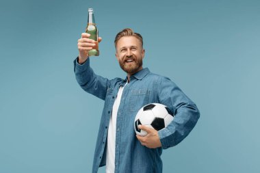 Yakışıklı, sakallı, hippi bir şişe bira ve klasik futbol topunu stüdyoda mavi arka planda tutuyor..