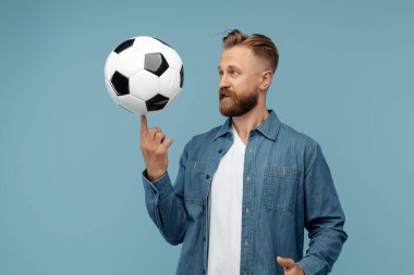 Neşeli sakallı sarışın hippi klasik futbol topunu stüdyoda mavi arka planda döndürür.