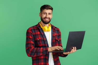 Yakışıklı sakallı hippi genç adam yeşil arka planda izole edilmiş bilgisayar kullanıyor. Kameraya bakıyorum..