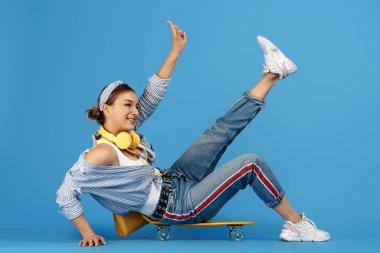 Kaygısız, sarı kulaklıklı genç bir kadın mavi arka planda kaykayın üzerinde oturuyor. Barış jesti gösteriyor.