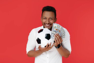 Etkileyici Afrikalı Amerikalı genç adam elinde nakit para ve kırmızı arka planda futbol topu tutuyor.