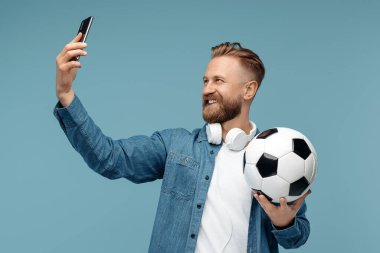 Yakışıklı sakallı hippi sarışın genç adam mavi arka planda klasik futbol topu tutarak akıllı telefondan selfie çekiyor..