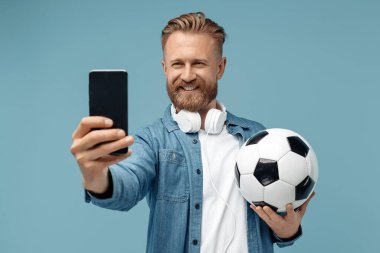 Neşeli sakallı hippi sarışın genç adam mavi arka planda klasik futbol topu tutarak akıllı telefondan selfie çekiyor..