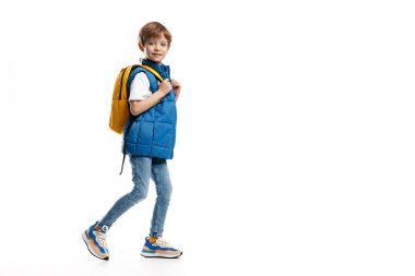 Sırt çantalı beyaz arka planda yürüyen küçük bir çocuğun tam boy görüntüsü. Okula geri dön. Kameraya bakıyorum..