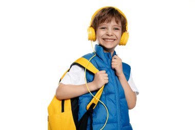 Kulaklık takan, sarı çantalı, beyaz arka planda kameraya bakan gülen küçük bir çocuk. Okula Dönüş.