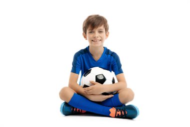 Mavi spor kıyafetli gülümseyen genç çocuk beyaz arka planda klasik futbol topuyla poz veriyor..