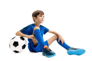 Mavi spor kıyafetli genç beyaz arka planda yerde klasik futbol topuyla poz veriyor..