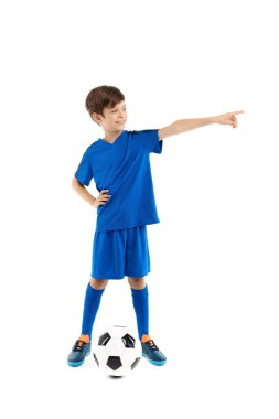 Mavi spor kıyafetli neşeli bir genç klasik futbol topuyla beyaz arka planı işaret ederek poz veriyor..