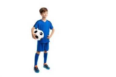 Mavi spor kıyafetli, beyaz arka planda klasik futbol topuyla poz veren kendinden emin genç bir çocuk..