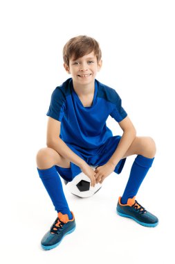 Mavi spor kıyafetli gülümseyen genç beyaz arka planda klasik futbol topunun üzerinde oturuyor..