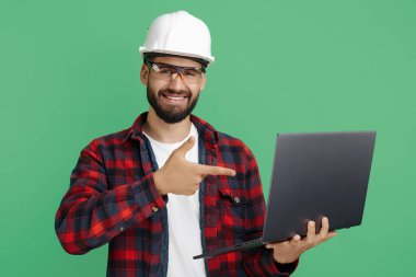 Sakallı mühendis ya da inşaatçı yeşil arka planda bilgisayarı işaret ediyor..