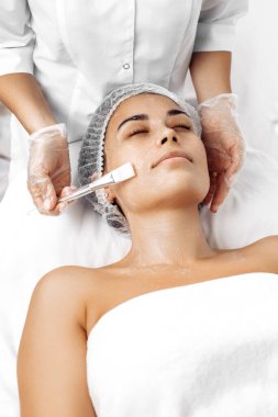 Kozmetik uzmanı kadın yüzüne kozmetoloji salonunda fırçayla krem sürüyor.
