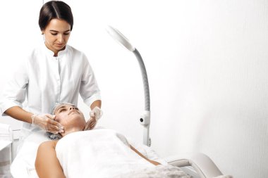 Kozmetolog kozmetoloji salonunda kadın yüzüne krem sürüyor.