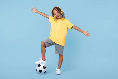 Afrikalı, kulaklıklı, bir ayağı futbol topunda mavi arka planda olan şık küçük bir çocuk..