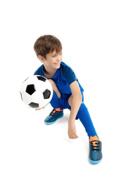 Mavi spor kıyafetli, yerde klasik futbol topuyla poz veren sevimli genç çocuk..