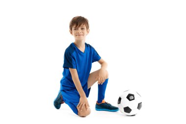 Mavi spor kıyafetli sevimli çocuk beyaz arka planda yerde klasik futbol topuyla poz veriyor..