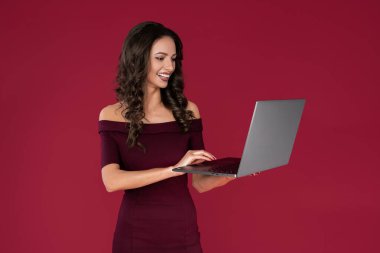 Kestane rengi elbise giymiş mutlu zarif genç bir kadın kırmızı arka planda bilgisayar ya da defter üzerinde parmakla işaret ediyor.