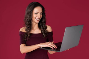 Kestane rengi elbise giymiş neşeli genç bir kadın kırmızı arka planda bilgisayar ya da not defterine işaret ediyor..