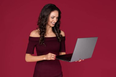 Kestane rengi elbise giymiş gülümseyen genç bir kadın kırmızı arka planda parmakla işaret ediyor..