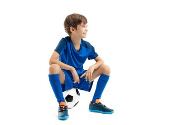 Mavi spor kıyafetli gülümseyen genç beyaz arka planda klasik futbol topunun üzerinde oturuyor..