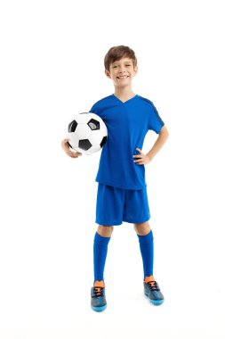 Mavi spor kıyafetli, beyaz arka planda klasik futbol topuyla poz veren kendinden emin genç bir çocuk..