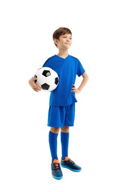 Mavi spor kıyafetli kendinden emin çocuk beyaz arka planda klasik futbol topuyla poz veriyor..