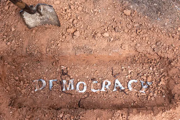 stock image Wood letters that spell out the word democracy, lie in a shallow grave with a shovel nearby, suggesting democracy is being buried.